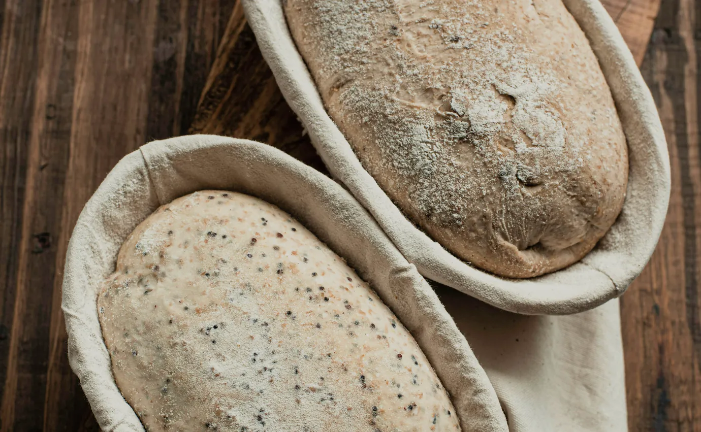 Two loaves rising in cane baskets on a dark bench