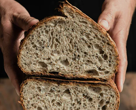 Two hands holding a halved country loaf, its open crumb towards the camera, against black