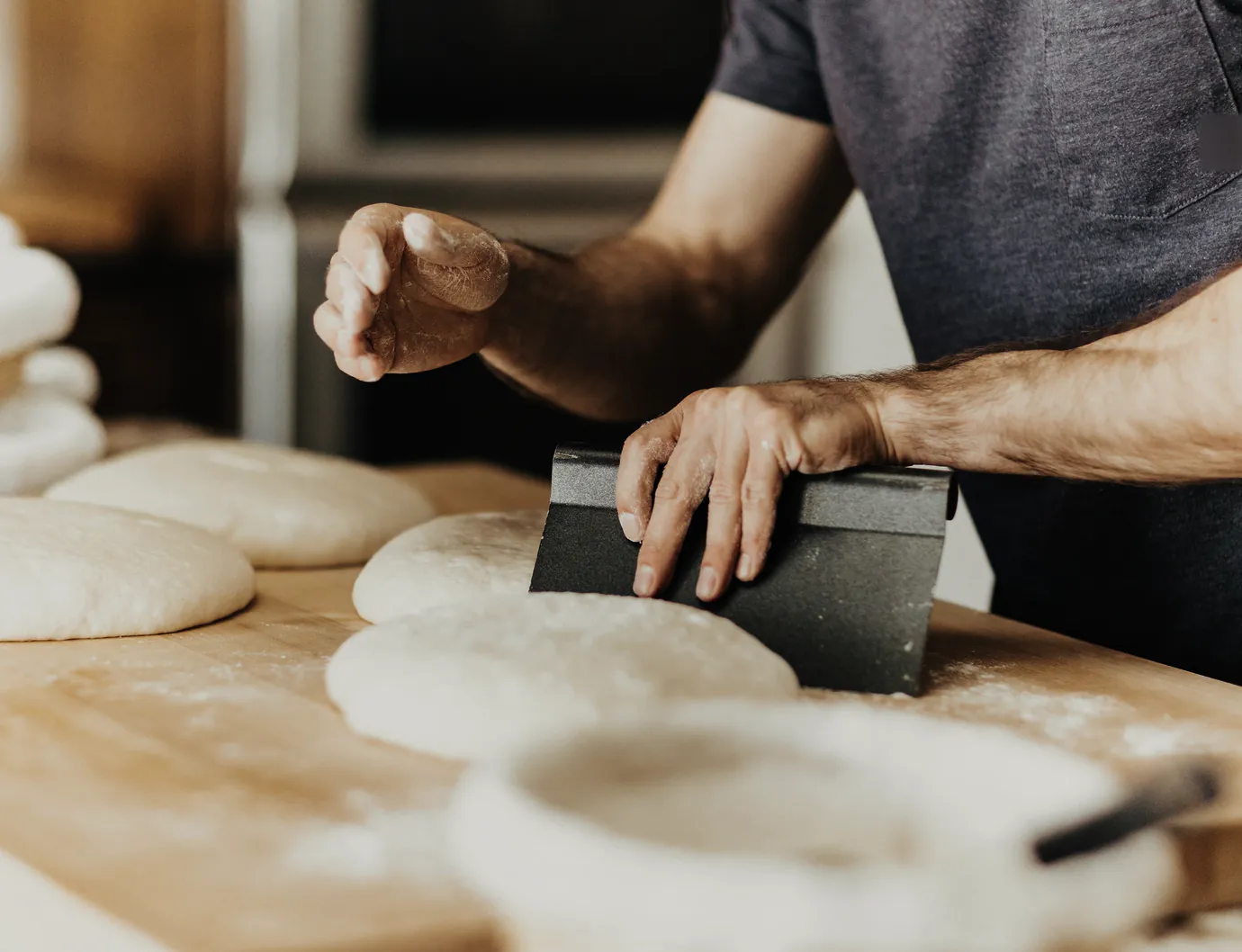A baker dividing dough with a bench scraper, rounds of dough along the board